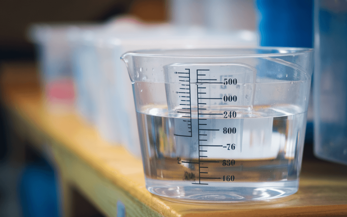 Un récipient mesurant de l'eau, marqué en millilitres, posé sur une table en bois, avec d'autres contenants flous en arrière-plan.