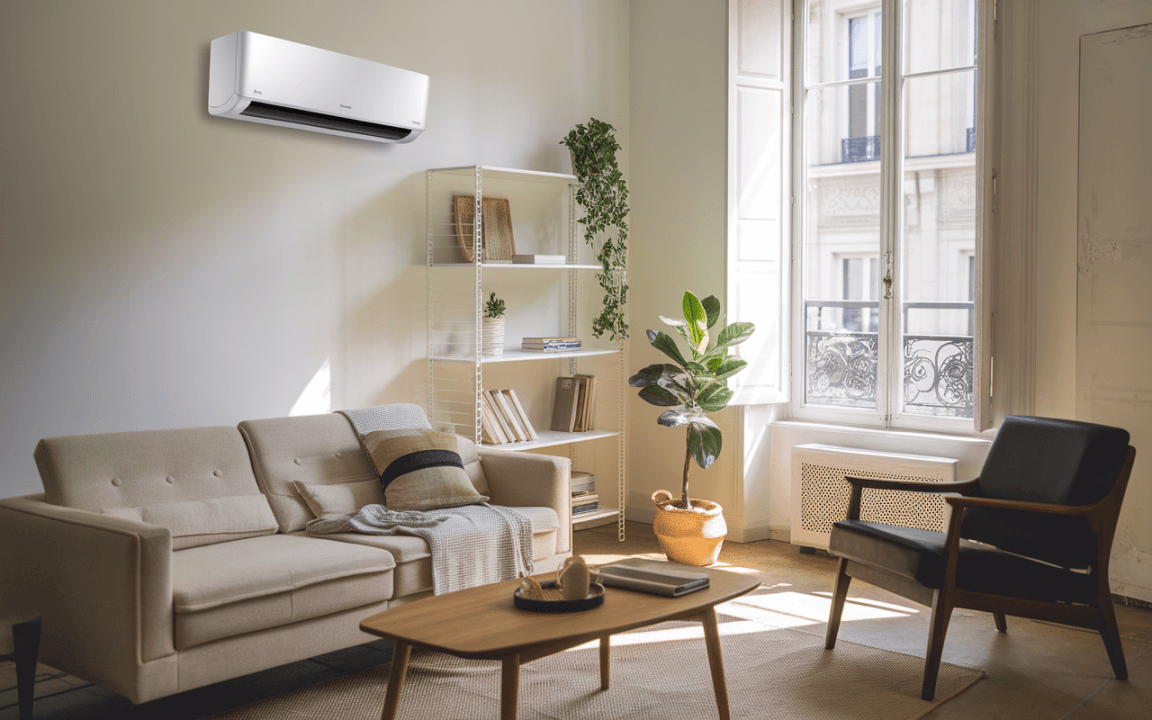 Intérieur lumineux avec un canapé beige, une table basse, des étagères, une plante verte, et une climatisation murale. Vue sur des fenêtres.