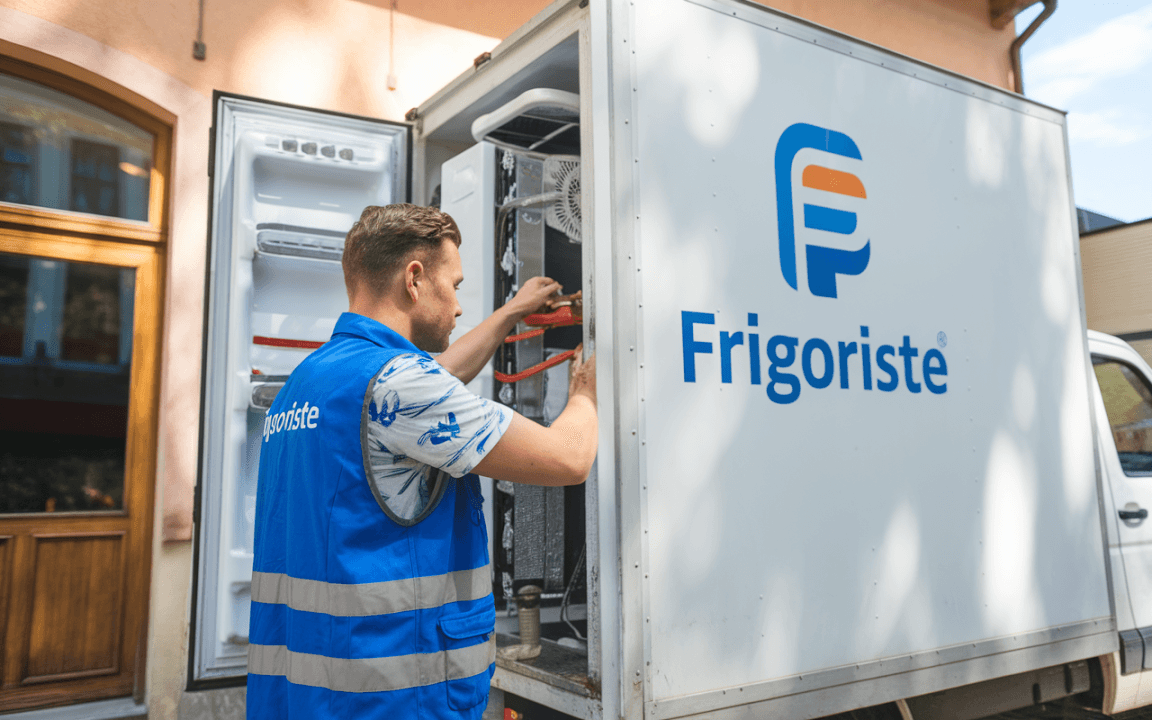 Technicien travaillant sur un réfrigérateur dans un véhicule Frigoriste, portant un gilet bleu avec logo. Éclairage naturel.