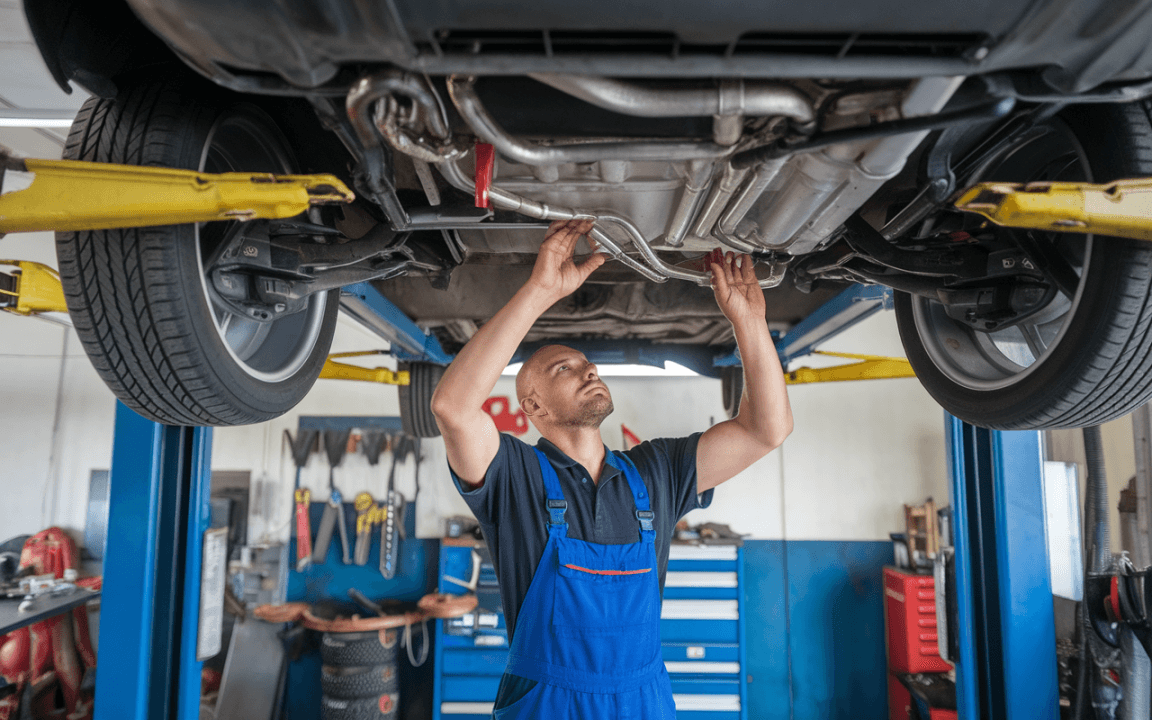 Un mécanicien inspecte le dessous d'une voiture levée dans un garage, utilisant des outils variés et en portant une combinaison bleue.