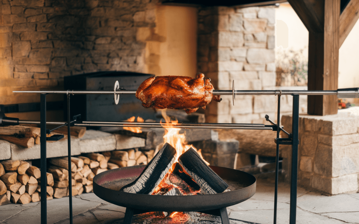 Un poulet rôti tourne sur une broche en fer, au-dessus d'un feu de bois dans un espace extérieur couvert.