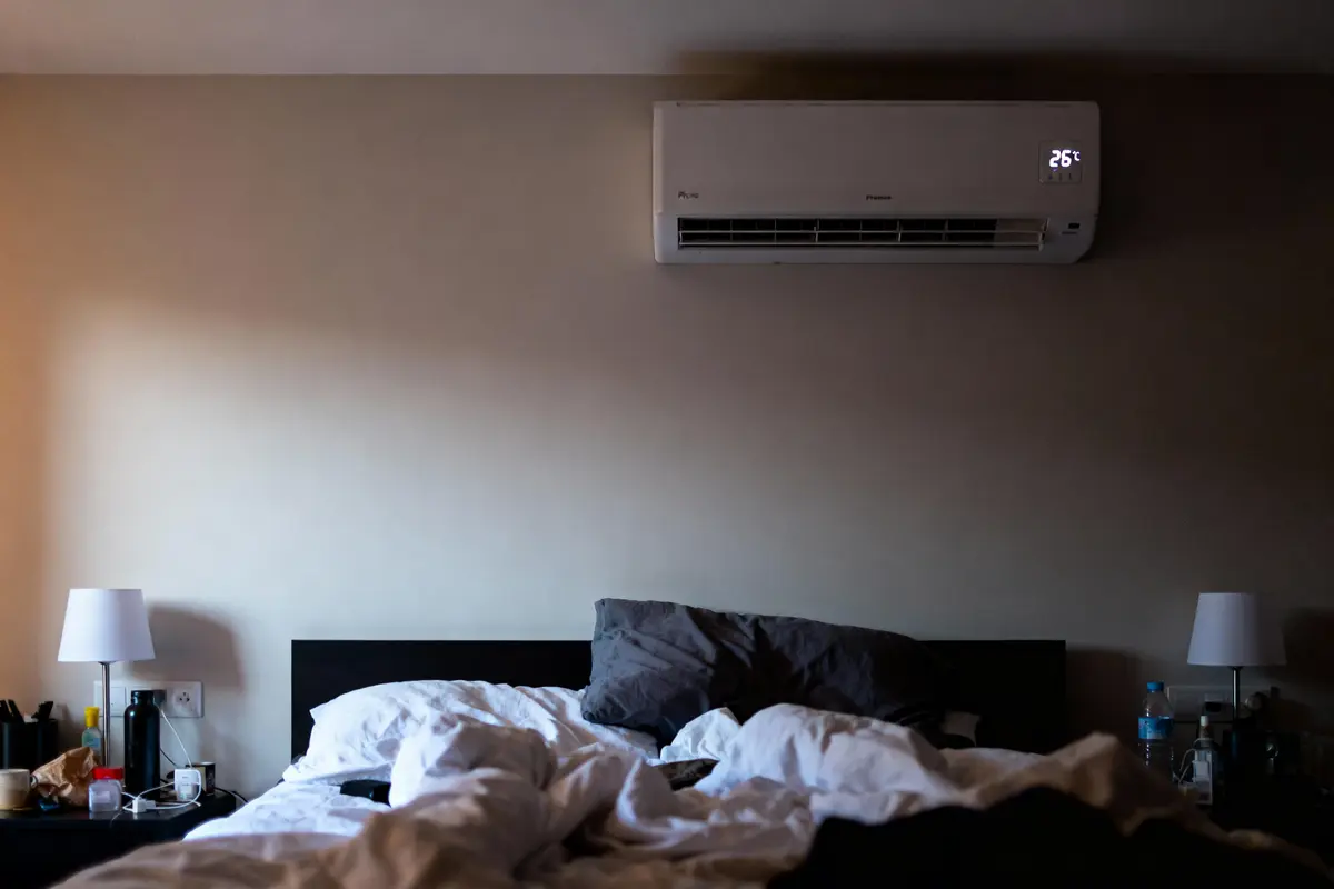 Chambre avec lit défait, lampes de chevet, climatiseur mural affichant 26°C, ambiance tamisée.