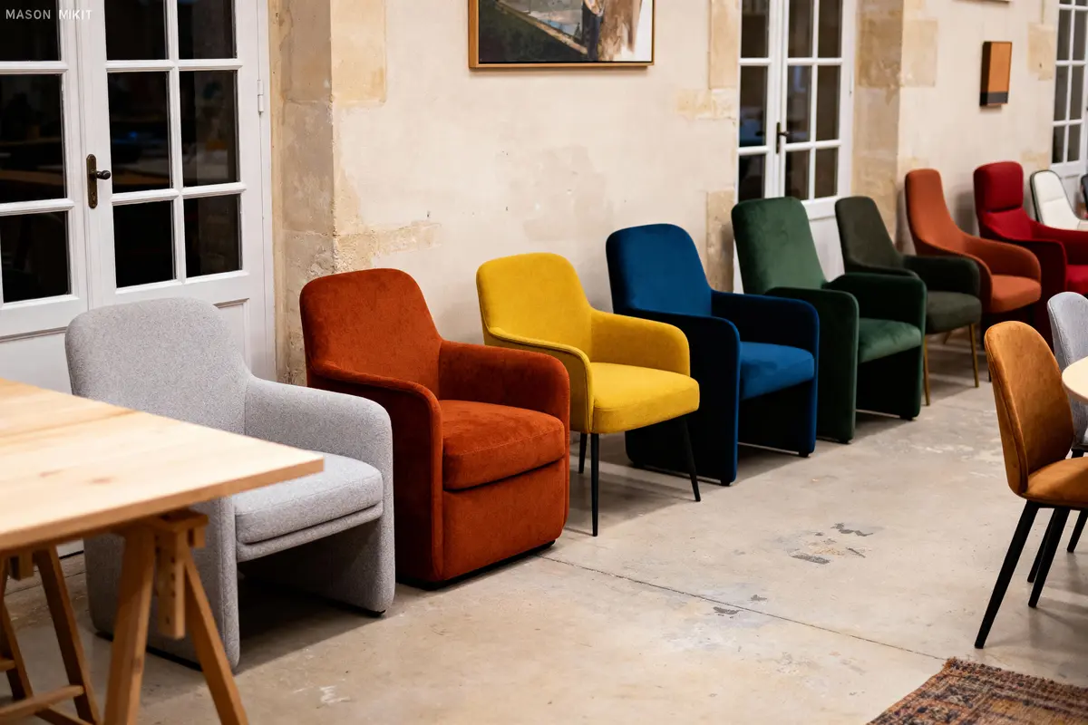 Six fauteuils colorés alignés contre un mur beige, devant une table en bois, ambiance intérieure chaleureuse.