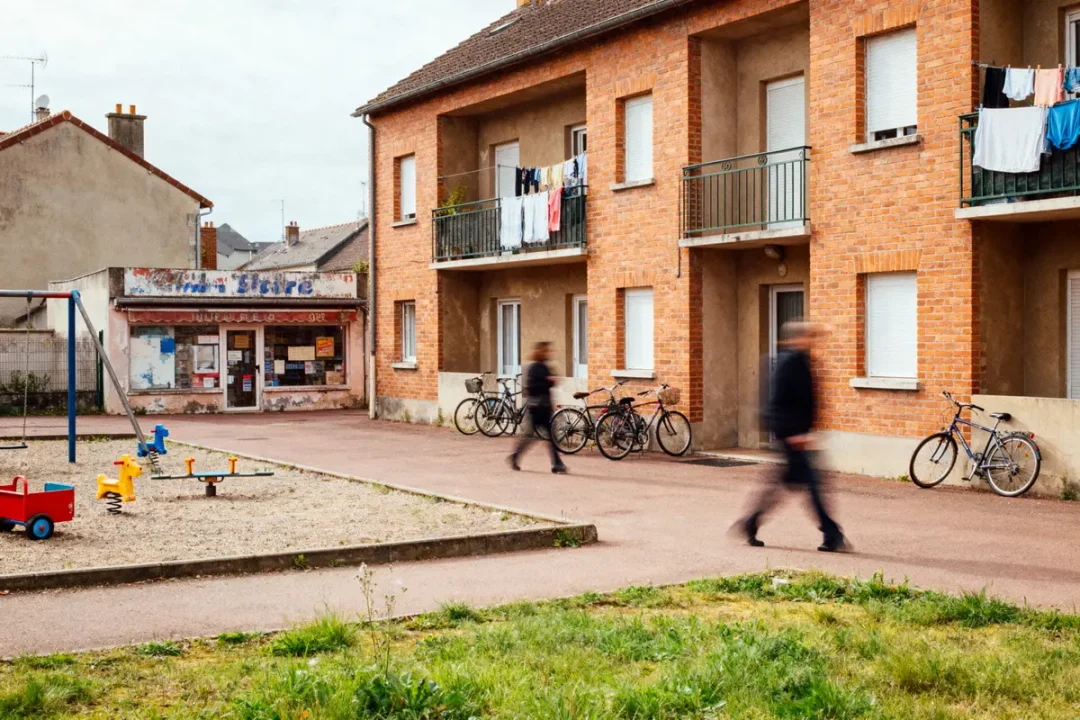 Immeuble en briques, balcons avec linge, vélos garés, deux personnes floues marchant, magasin au fond, aire de jeux.