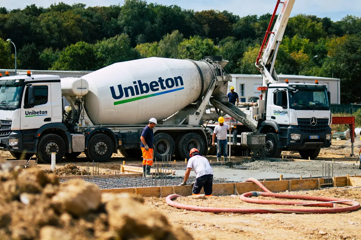Camions-toupies Unibeton sur chantier, ouvriers coulant du béton, environnement forestier en arrière-plan.