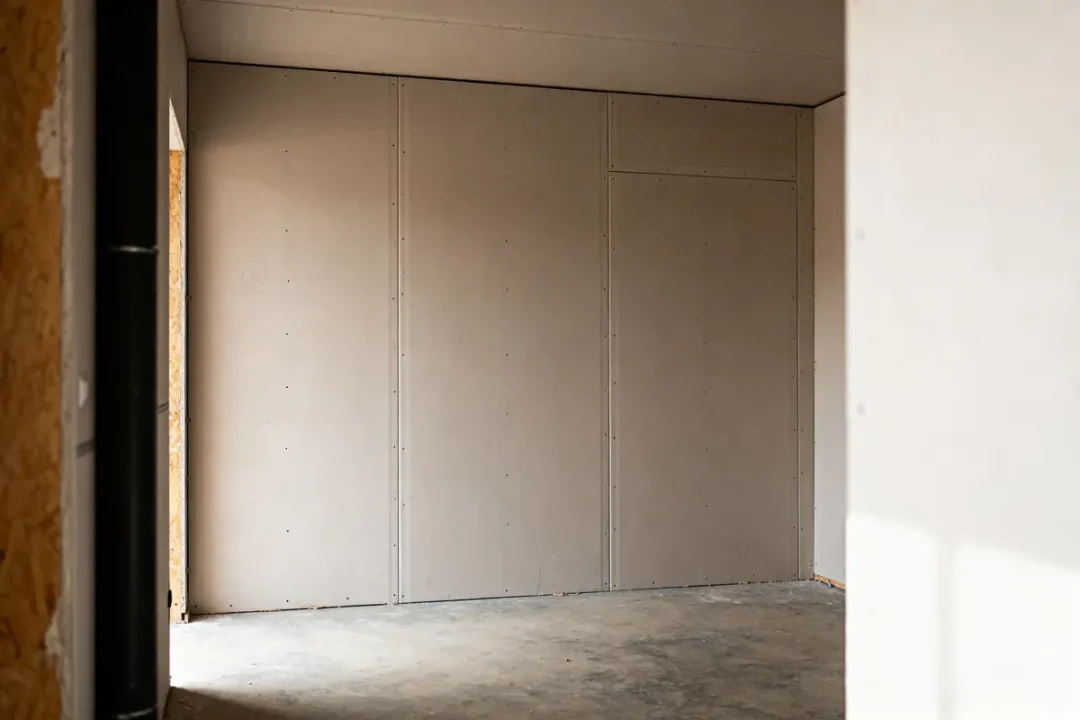 Pièce vide avec sol en béton, murs en plaques de plâtre, éclairage naturel venant de la gauche.