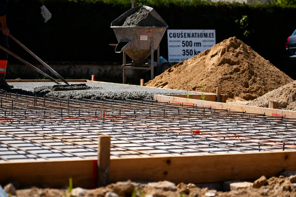 Ouvrier coule du béton sur armature métallique, chantier avec sable et bétonnière en arrière-plan.