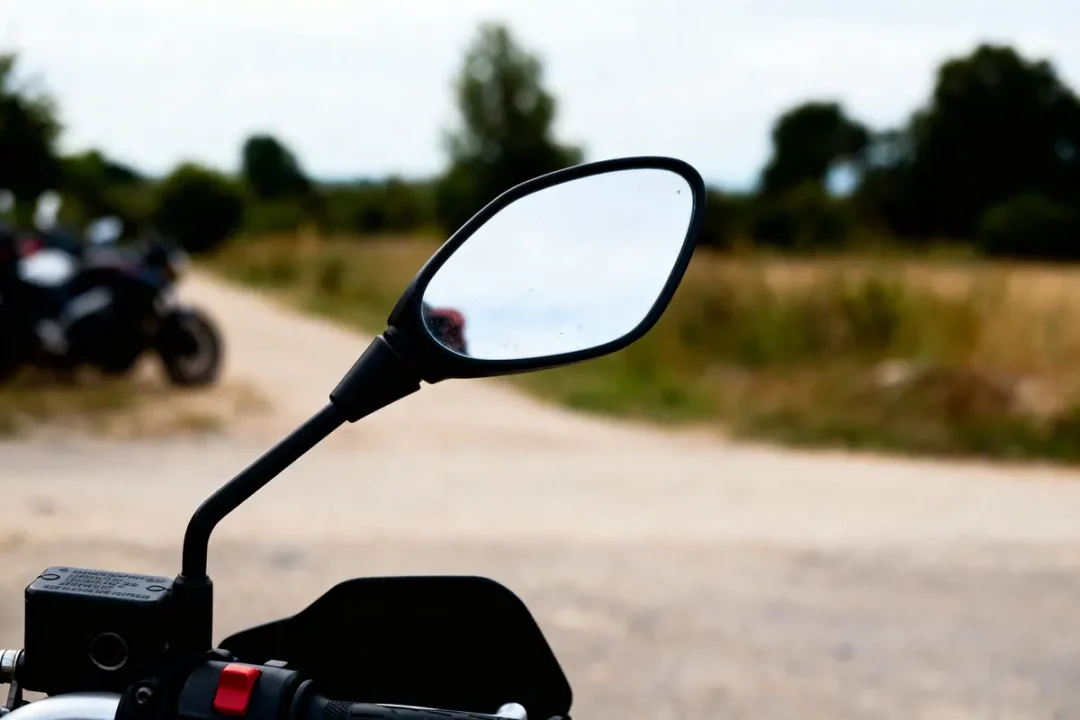 Rétroviseur de moto reflétant paysage flou avec route en arrière-plan, ciel légèrement nuageux.