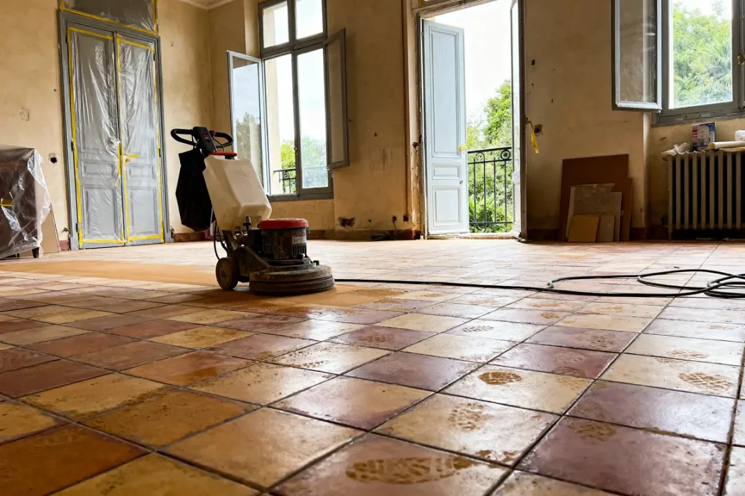 Pièce vide avec sol en tomettes, fenêtres ouvertes, machine de rénovation de sol au premier plan.