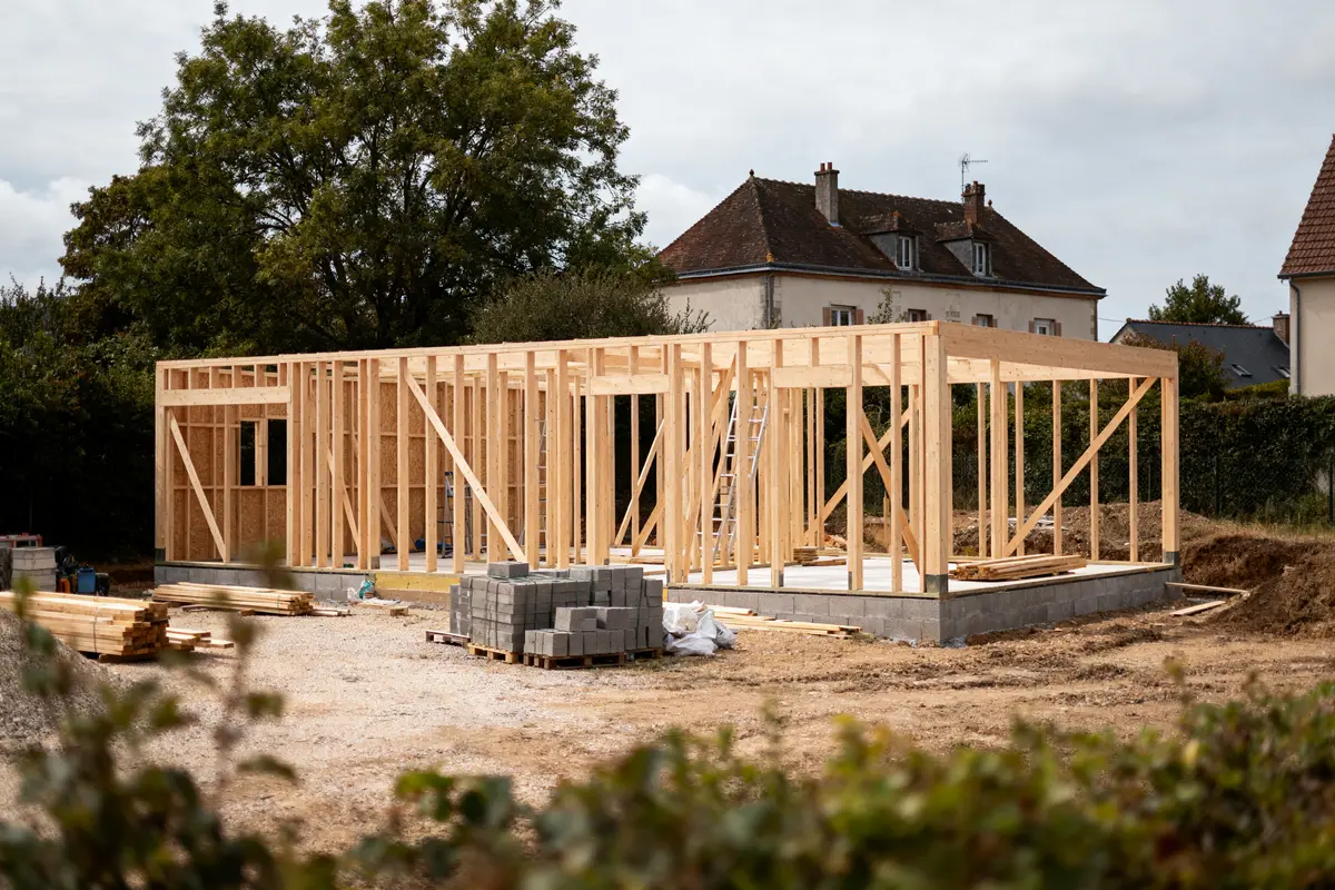 Structure en bois en construction avec maison et arbres en arrière-plan, divers matériaux au sol.