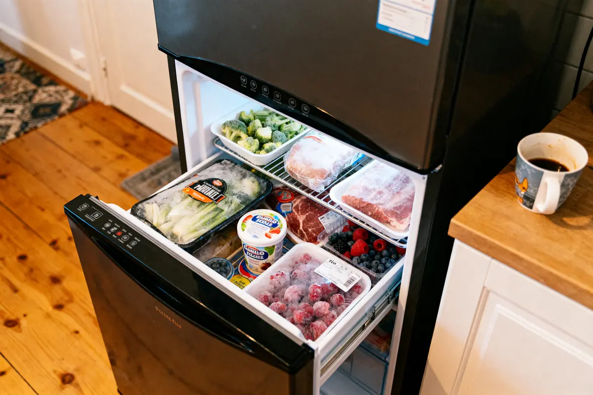 Congélateur ouvert rempli d'aliments surgelés, tels que légumes, viandes, fruits rouges, avec tasse de café à côté.
