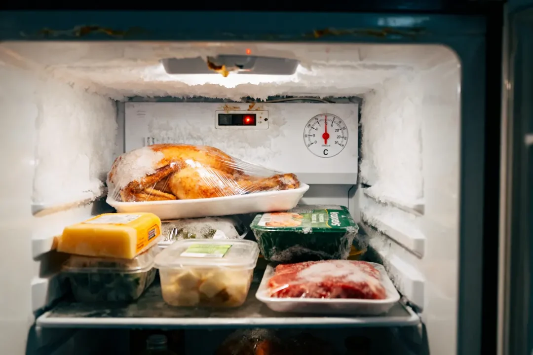 Congélateur ouvert avec aliments congelés, incluant poulet, fromage, viandes, emballés sous film plastique. Thermomètre visible.