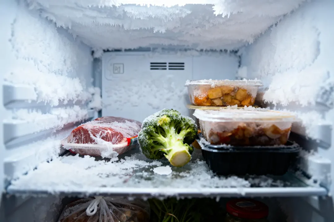 Congélateur rempli de givre contenant aliments congelés : viande, brocoli, plats préparés empaquetés.