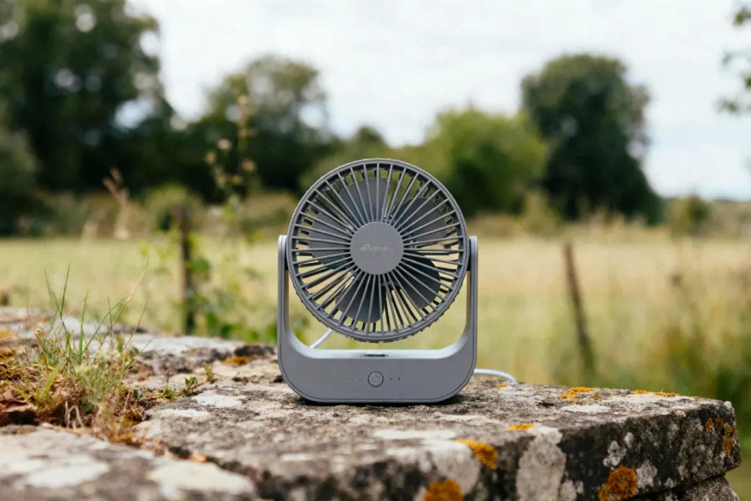 Petit ventilateur portable gris sur un muret en pierre, paysage verdoyant en arrière-plan.
